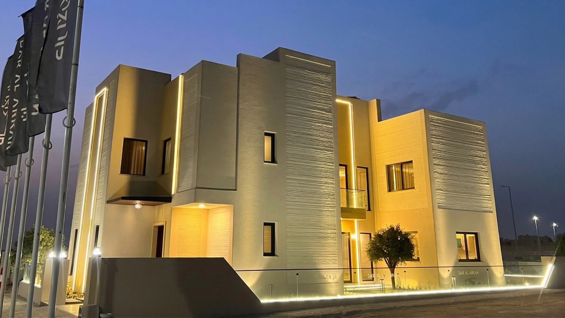 Front side view of a 3 storey cream coloured 3d printed concrete house lit up by lights in the evening