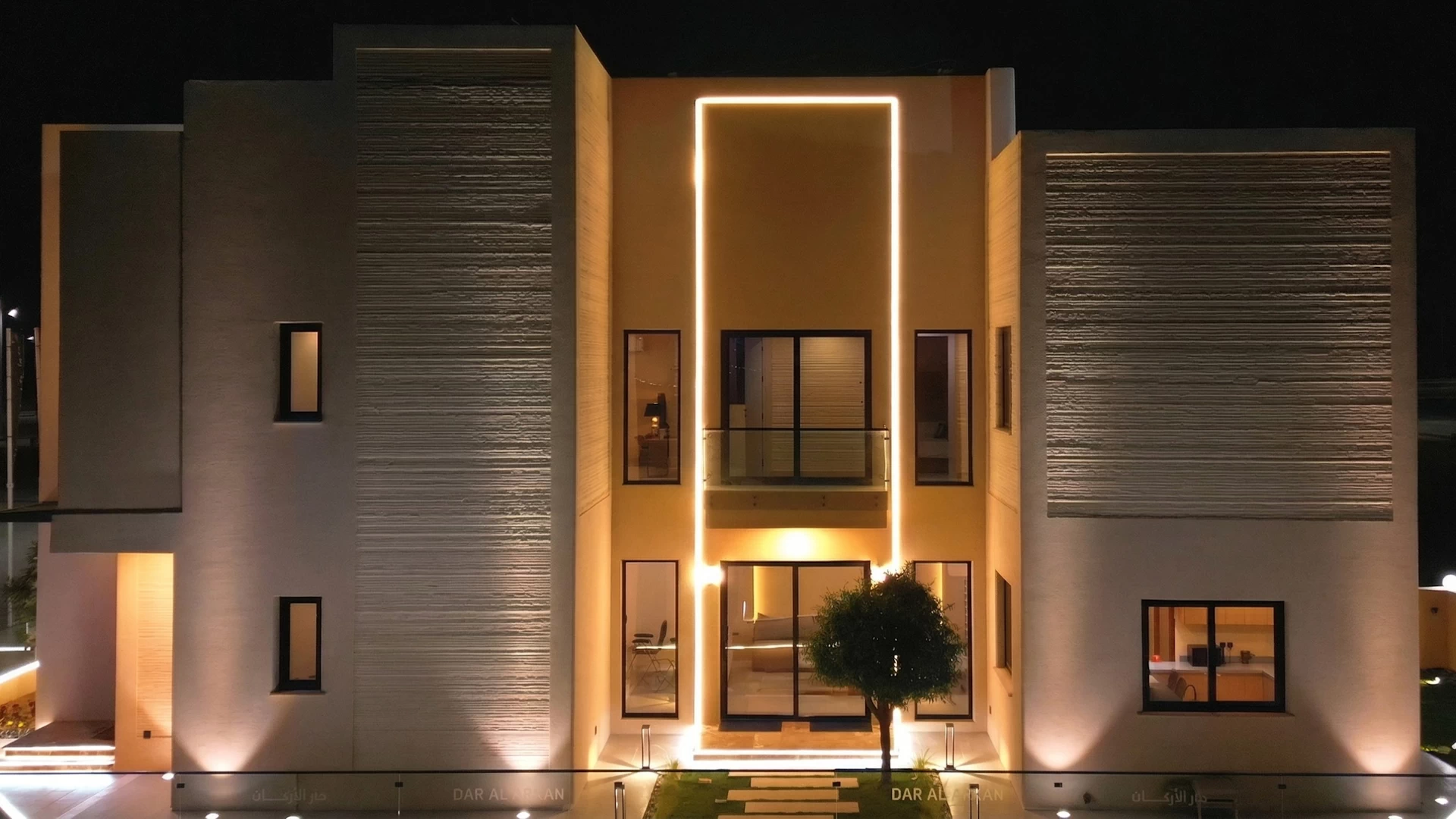 Front view of a 3 storey cream coloured 3d printed concrete house lit up by lights in the night