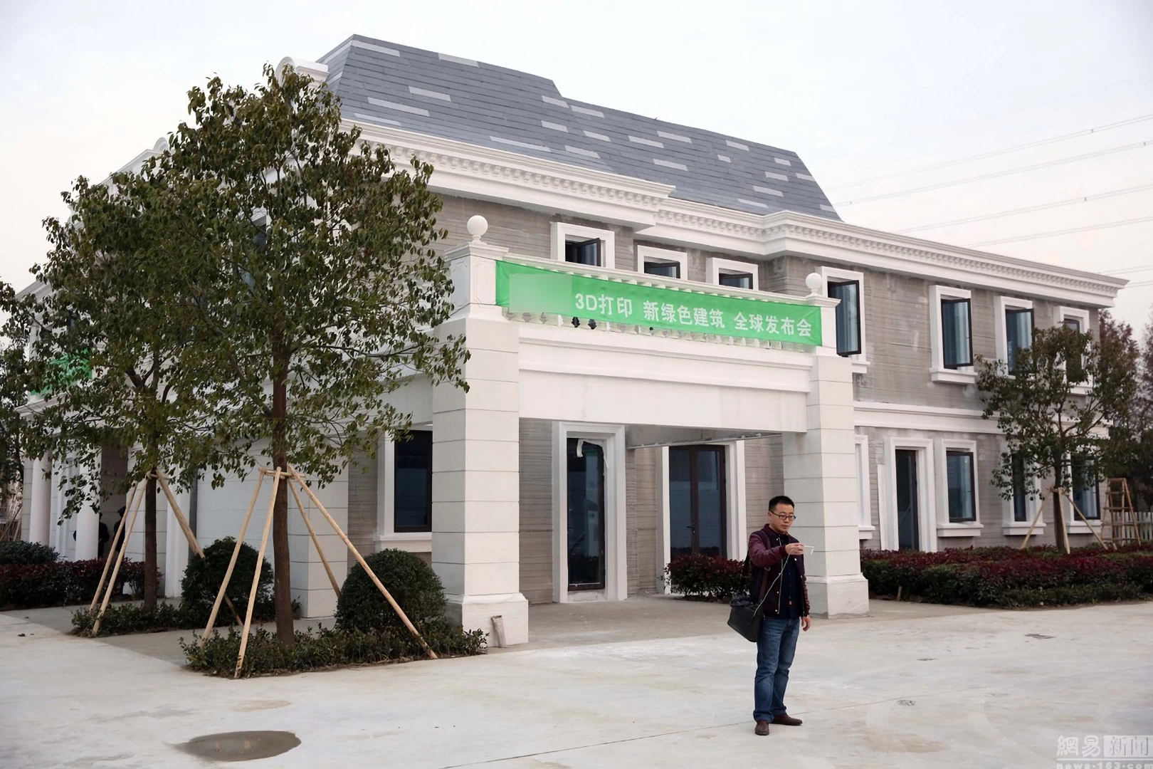 Rear view of white 3d printed double storey concrete house with grey roof and surrounded by plants