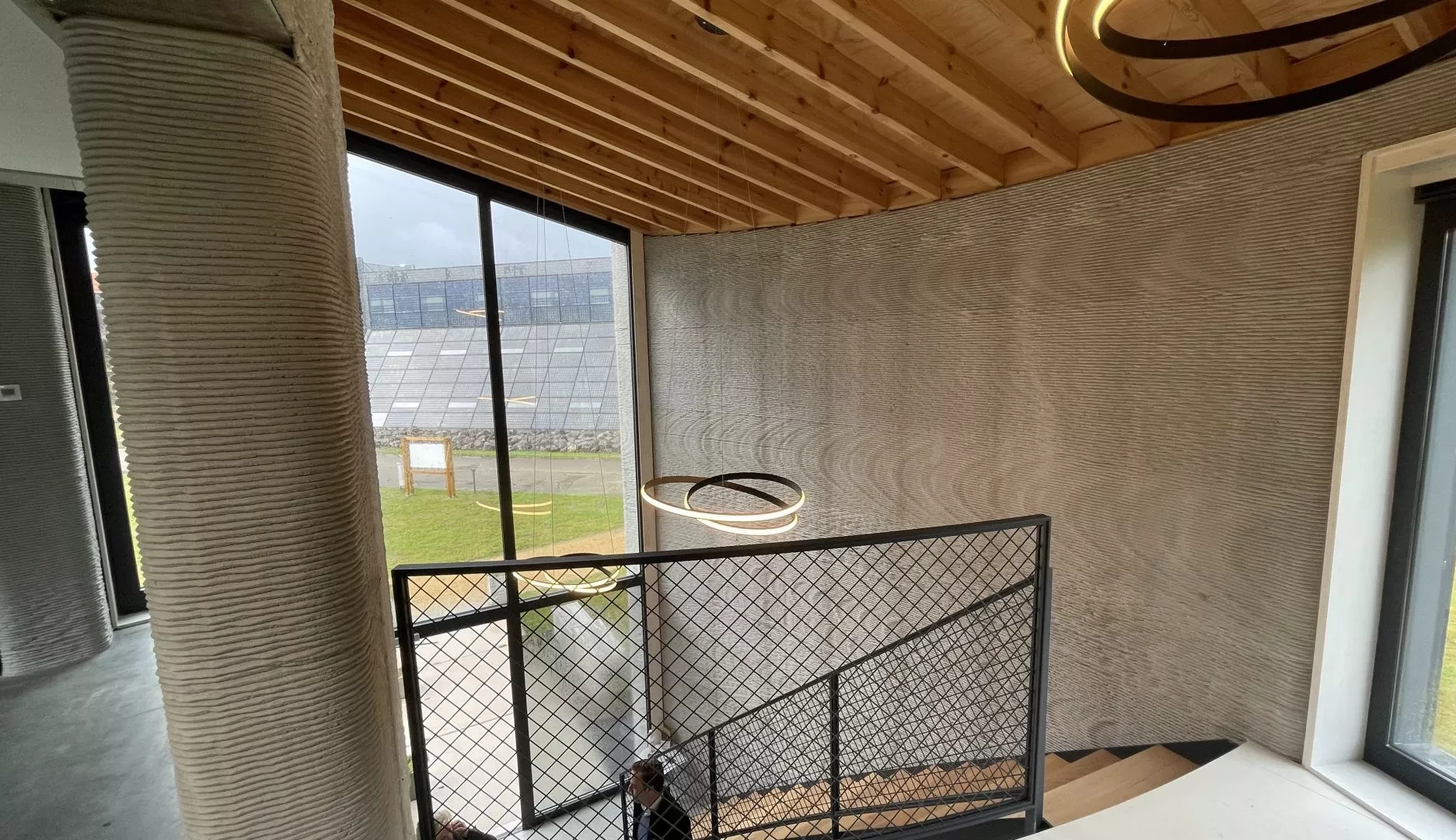 View of stairway inside 3d printed concrete house with large window and wooden ceiling beams