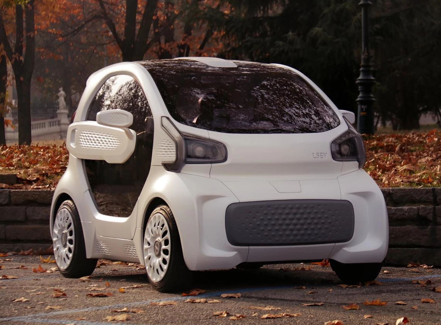 Front right view of small white 3d printed plastic 2 seater car next to a park in autumn