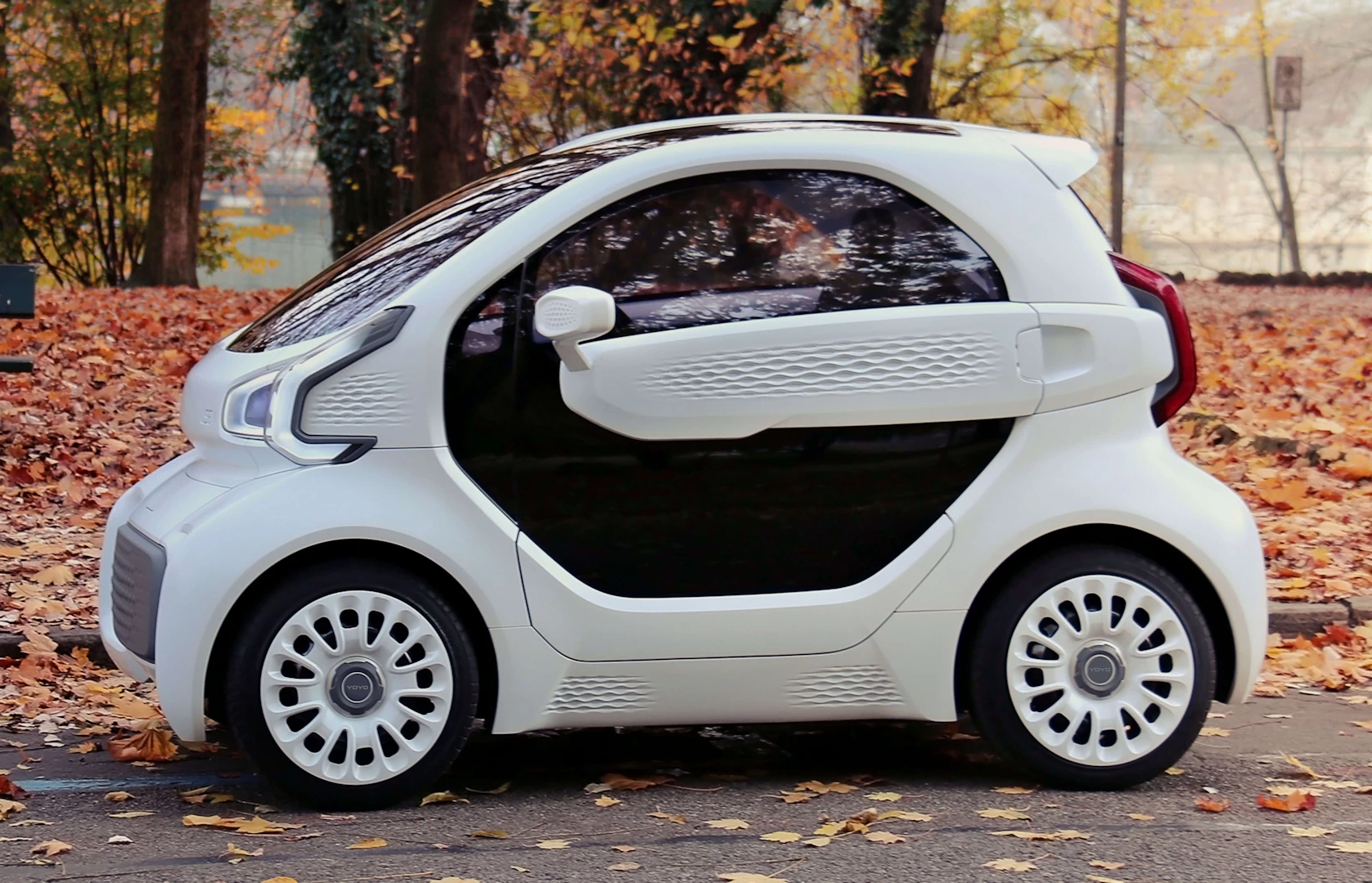 Left view of small white 3d printed plastic 2 seater car next to a park in autumn